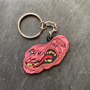Op de afbeelding: Een roze rubberen sleutelhanger met een cartoonmonster met een wijd open mond en scherpe tanden. De sleutelhanger is bevestigd aan een zilveren metalen ring en ketting.