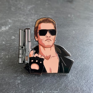 Puede incluir: Un pin de esmalte plateado y negro con el retrato de un hombre con gafas de sol y una chaqueta de cuero, sosteniendo una pistola. El hombre tiene el pelo corto castaño y una expresión seria.