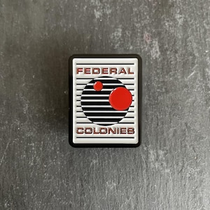 May include: A black and white enamel pin with the text "FEDERAL COLONIES" in red lettering. The pin features a black circle with white stripes and two red circles inside.