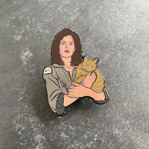 Puede incluir: Un pin de esmalte en blanco y negro que representa a una mujer con cabello castaño sosteniendo un gato jengibre. La mujer lleva una chaqueta oscura con un parche en el hombro. El pin es un personaje popular de la película Alien.