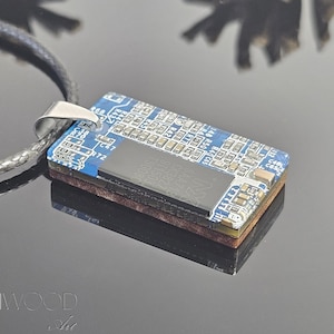 May include: A rectangular pendant made from a blue circuit board, featuring silver metal hardware and a black cord. The pendant has a dark brown layer and a black component on top. The pendant is approximately 1.5 inches long.