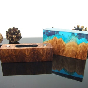 May include: Two wooden boxes. One is a rectangular jewelry box with a black insert and silver accents. The other is a rectangular box with a wood grain base and a blue and turquoise resin top, resembling a landscape. Pine cones are in the background.