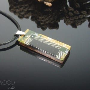 May include: A rectangular pendant necklace featuring a green circuit board component with visible metallic pins and a black central panel, attached to a black cord by a silver-toned metal bail. The text TRINWOOD Art is visible in the corner.