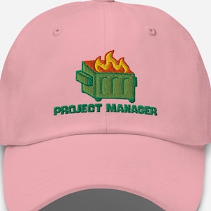 May include: A light pink baseball cap with an embroidered design. The design features a green dumpster on fire with yellow and orange flames, and the words "PROJECT MANAGER" in green.