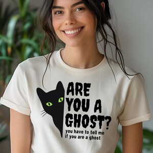 May include: Cream-colored t-shirt featuring a black cat graphic with green eyes and the text "ARE YOU A GHOST? you have to tell me if you are a ghost." The shirt has short sleeves and a crew neck.