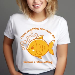 Op de afbeelding: Wit T-shirt met een oranje cartoonvis en bellen ontwerp. De tekst luidt: "Ik leer elke dag iets nieuws omdat ik niets onthoud". Het ontwerp is gecentreerd en biedt een humoristische stijl.