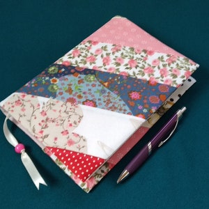 Puede incluir: Un cuaderno hecho a mano con una cubierta de patchwork colorida con motivos florales y geométricos. El cuaderno tiene un marcapáginas de cinta rosa y un bolígrafo morado.