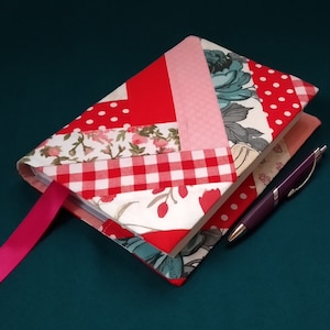 Puede incluir: Una cubierta de diario de tela roja, rosa, blanca y azul con un patrón de cuadros vichy y un marcapáginas de cinta rosa. Un bolígrafo morado está sobre la cubierta.