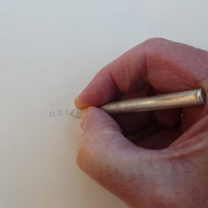 Könnte beinhalten: Eine Hand, die einen silberfarbenen Bleistift hält und die Buchstaben "abc" auf eine weiße Oberfläche schreibt. Der Bleistift hat eine zylindrische Form mit abgerundeter Spitze. Die Buchstaben sind hellgrau geschrieben.