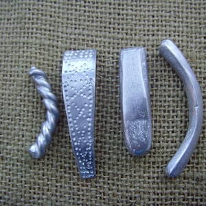 May include: Four silver metal objects are displayed on a textured, tan surface. The objects include a twisted rod, a flat piece with dotted patterns, a rectangular piece, and a curved piece. The objects appear to be handmade.