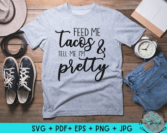 May include: Gray t-shirt with black text that reads "Feed me tacos & tell me I'm pretty". The shirt is laying on a wooden surface with other items around it.