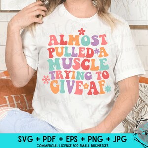 May include: White t-shirt with the phrase "ALMOST PULLED A MUSCLE TRYING TO GIVE AF" in colorful, retro-style lettering. The design includes flower accents. The bottom of the image shows file type options and a logo.