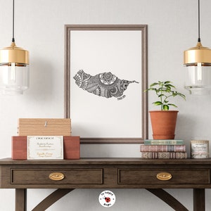 May include: A black and white line art print of the island of Grenada with intricate patterns. The print is framed in a light brown frame and is hanging on a white wall. The print is displayed on a shelf with a wooden box, a potted plant, and a stack of books.