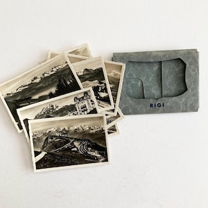 May include: A collection of vintage black and white postcards featuring mountain landscapes and architecture, with a gray card holder. The postcards have a deckled edge. The card holder is labeled "RIGI".