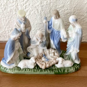 Könnte beinhalten: Ein Krippenfiguren-Set aus Porzellan mit fünf Figuren und zwei Lämmern auf einem grünen Sockel. Die Figuren tragen weiß-blaue Gewänder und umgeben eine zentrale Krippe mit einem Baby. Das Stück hat eine glänzende Oberfläche und Details.