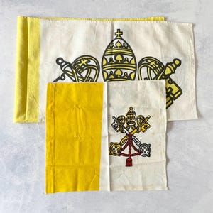 Puede incluir: Dos banderas rectangulares de tela con el emblema de la Ciudad del Vaticano. La bandera más grande muestra una franja vertical amarilla y un campo blanco con una tiara papal y llaves cruzadas negras. La bandera más pequeña tiene una franja amarilla y un campo blanco con una tiara papal y llaves de colores.