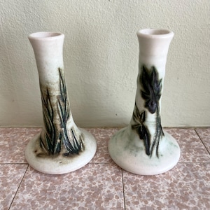 Op de afbeelding: Twee keramische vaasjes met een witte basis en een uitlopende bovenkant. Eén vaas heeft een groen en bruin plantenontwerp, de andere een vergelijkbaar ontwerp. Deze vazen zijn geschikt voor het uitstallen van kleine bloemen.