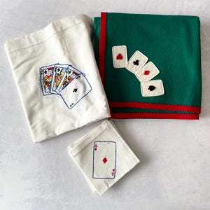 May include: A set of linens featuring playing card motifs. One off-white cloth displays an embroidered hand of cards. A green cloth with red trim features four felt card appliqués. A small off-white napkin shows an embroidered ace of diamonds.