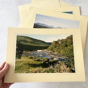 May include: A stack of rectangular paper prints featuring landscape photography. The top print displays a river flowing through a green valley with hills in the background, framed by a cream-colored border with thin black lines.