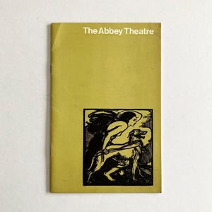 以下が含まれることがあります： アビーシアターのヴィンテージパンフレット。マスタードイエローの表紙に「The Abbey Theatre」の文字が白で書かれています。女性と犬の白黒イラストが前面にあります。