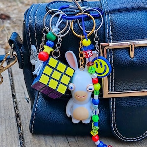 May include: A dark blue textured handbag featuring a blue carabiner clip holding two keychains. One keychain has a miniature yellow, red, and blue puzzle cube and a white butterfly charm. The other features a white rabbit figure, a yellow smiley face, and a long strand of colorful plastic beads with a red letter K cube.