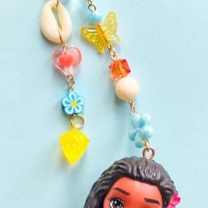 Puede incluir: Un colorido dije de cuentas con una mu&ntilde;eca de pelo largo negro y una flor rosa. El dije incluye una concha, un coraz&oacute;n, una flor, una mariposa y cuentas geom&eacute;tricas en amarillo, azul, naranja y blanco, todas conectadas por cadenas doradas.