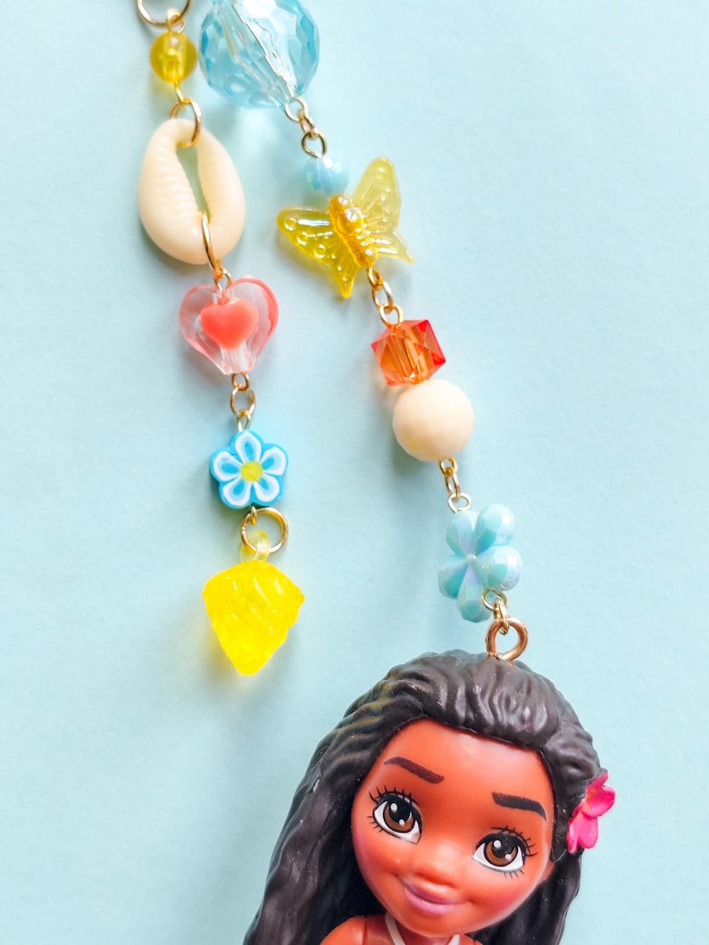 Puede incluir: Un colorido dije de cuentas con una mu&ntilde;eca de pelo largo negro y una flor rosa. El dije incluye una concha, un coraz&oacute;n, una flor, una mariposa y cuentas geom&eacute;tricas en amarillo, azul, naranja y blanco, todas conectadas por cadenas doradas.