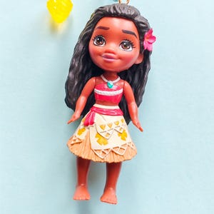Puede incluir: Peque&ntilde;a mu&ntilde;eca de un personaje de dibujos animados con cabello oscuro, que lleva una parte superior roja y blanca, una falda beige con detalles amarillos y una flor rosa en el pelo. La mu&ntilde;eca cuelga de una cadena de cuentas.