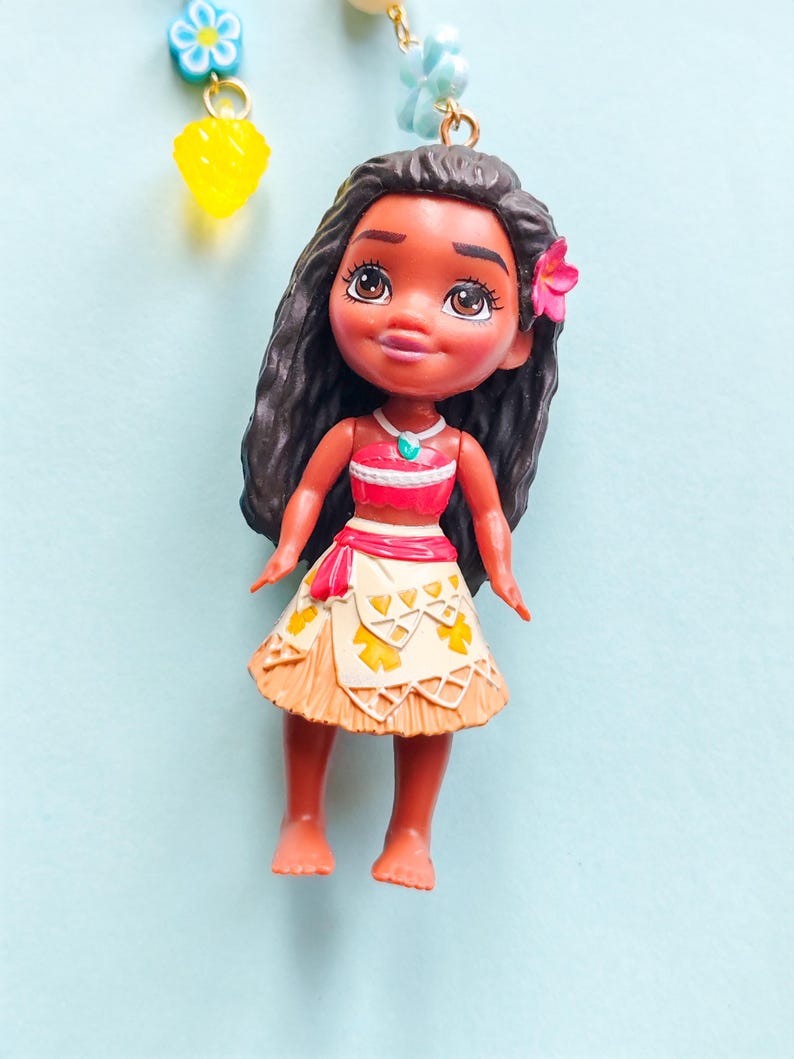 Puede incluir: Peque&ntilde;a mu&ntilde;eca de un personaje de dibujos animados con cabello oscuro, que lleva una parte superior roja y blanca, una falda beige con detalles amarillos y una flor rosa en el pelo. La mu&ntilde;eca cuelga de una cadena de cuentas.