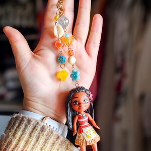 Puede incluir: Un llavero dorado con un dije que presenta una peque&ntilde;a mu&ntilde;eca y cuentas de colores. La mu&ntilde;eca tiene cabello oscuro y lleva una blusa roja y una falda. El dije incluye conchas marinas, flores y una mariposa.