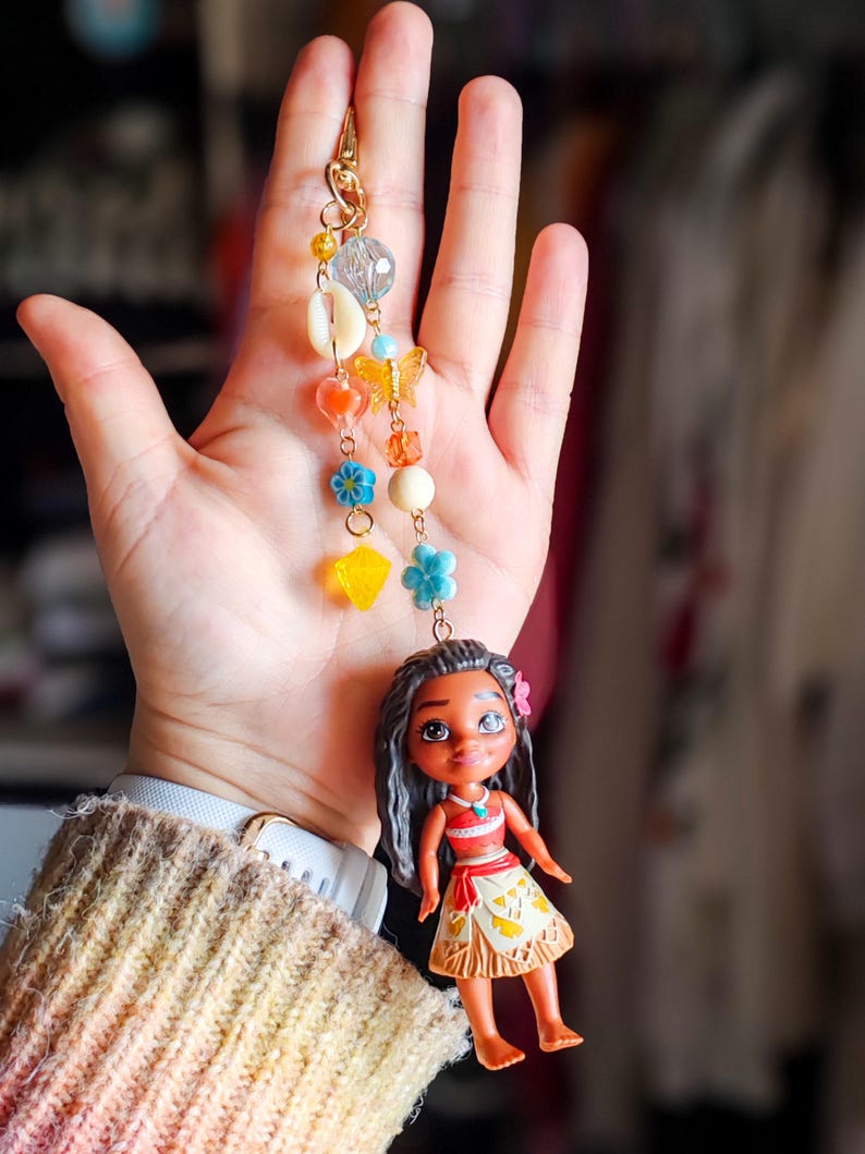 Puede incluir: Un llavero dorado con un dije que presenta una peque&ntilde;a mu&ntilde;eca y cuentas de colores. La mu&ntilde;eca tiene cabello oscuro y lleva una blusa roja y una falda. El dije incluye conchas marinas, flores y una mariposa.