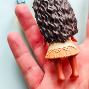 Puede incluir: Una peque&ntilde;a figura de un personaje con cabello rizado casta&ntilde;o oscuro, que lleva una falda beige y una parte superior blanca. La figura se sostiene en una mano, con un collar visible en el fondo.