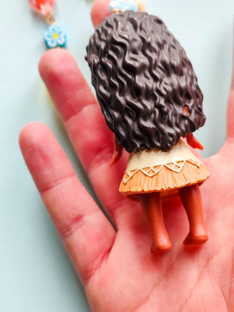 Puede incluir: Una peque&ntilde;a figura de un personaje con cabello rizado casta&ntilde;o oscuro, que lleva una falda beige y una parte superior blanca. La figura se sostiene en una mano, con un collar visible en el fondo.