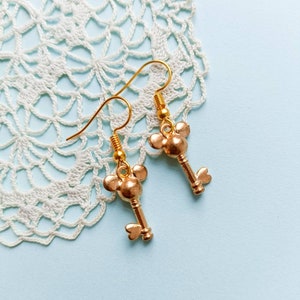 Könnte beinhalten: Goldene Schlüssel-Ohrringe mit einem Mickey-Maus-Kopf-Design. Die Ohrringe befinden sich auf einer weißen Häkelspitze.