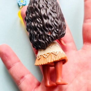 Puede incluir: Una peque&ntilde;a mu&ntilde;eca con cabello casta&ntilde;o oscuro y rizado, que lleva una falda beige y naranja. La mu&ntilde;eca forma parte de un collar con cuentas de colores y detalles florales. La mu&ntilde;eca est&aacute; en la mano de una persona.