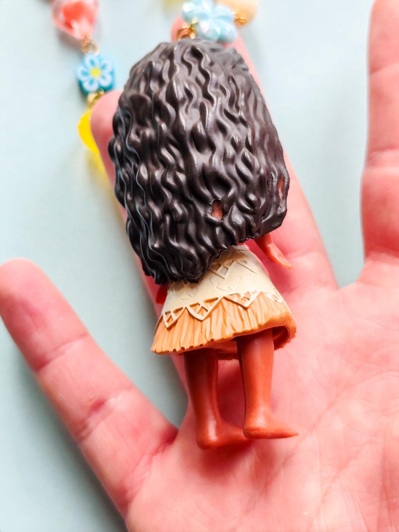 Puede incluir: Una peque&ntilde;a mu&ntilde;eca con cabello casta&ntilde;o oscuro y rizado, que lleva una falda beige y naranja. La mu&ntilde;eca forma parte de un collar con cuentas de colores y detalles florales. La mu&ntilde;eca est&aacute; en la mano de una persona.