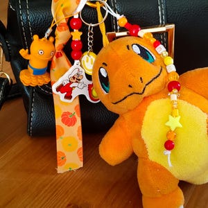 May include: A keychain accessory featuring an orange plush Charmander character, a yellow smiley face charm, a Mario character cutout, and an orange ribbon with a pumpkin pattern. The design includes red, yellow, and white beads on a metal ring.
