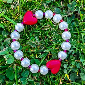 May include: A bracelet made of large, round, white faux pearls and two red heart-shaped beads. The bracelet is strung on a purple cord. The image is set against a background of green grass.