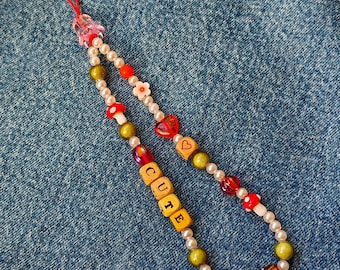 May include: A phone charm with a red string, white and green beads, red and white flowers, red mushrooms, a red heart, and wooden letter beads spelling "CUTE".