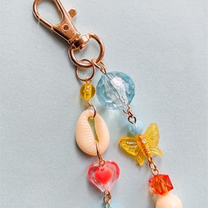 Puede incluir: Un llavero decorativo con un cierre dorado y un dije con una concha, una mariposa, un coraz&oacute;n y una flor. El dije tambi&eacute;n incluye varias cuentas coloridas en azul, amarillo, naranja y melocot&oacute;n.