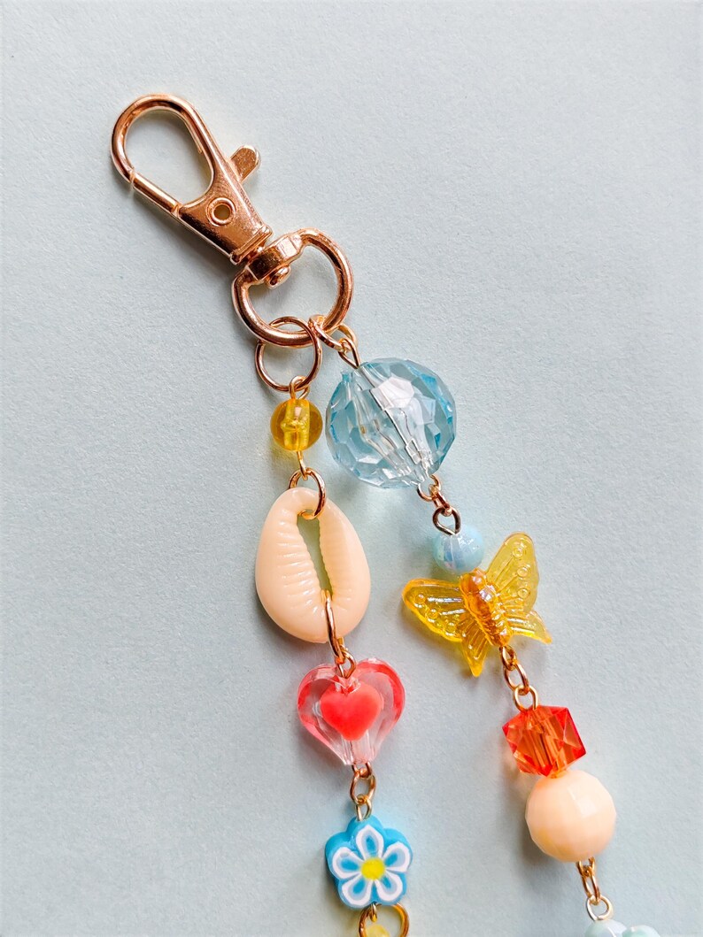 Puede incluir: Un llavero decorativo con un cierre dorado y un dije con una concha, una mariposa, un coraz&oacute;n y una flor. El dije tambi&eacute;n incluye varias cuentas coloridas en azul, amarillo, naranja y melocot&oacute;n.