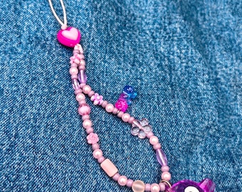 May include: A pink and purple beaded phone charm with a clear purple pig charm. The charm is on a denim background.