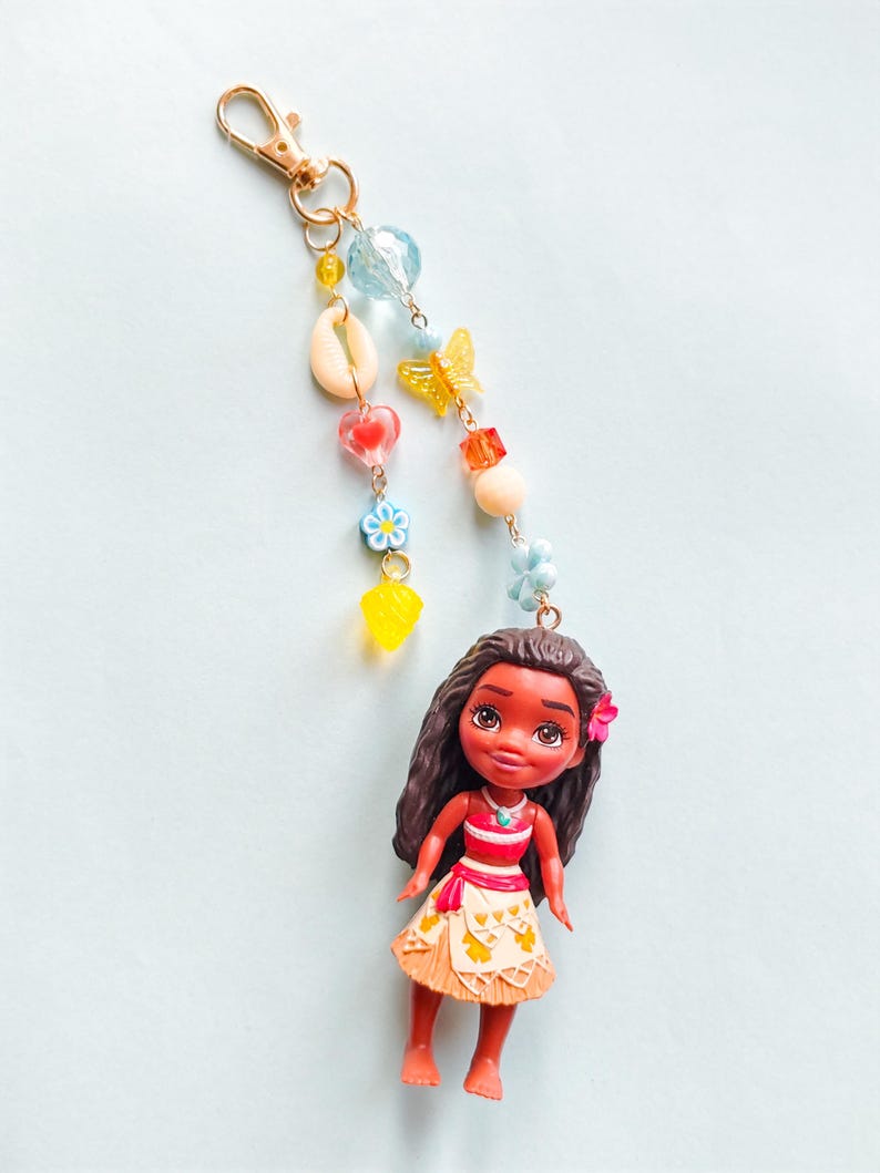 Puede incluir: Un llavero de mu&ntilde;eca con una peque&ntilde;a mu&ntilde;eca de pelo largo y oscuro, con una camiseta roja y blanca y una falda estampada. El llavero incluye un cierre dorado y una cadena de cuentas, conchas y abalorios de colores, incluyendo una mariposa y flores.
