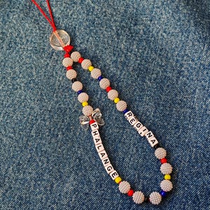 Puede incluir: Un dije de teléfono con cuentas con el nombre "PHALANGE REGINA" escrito con cuentas de letras blancas. El dije presenta cuentas texturizadas de color plateado y cuentas de acento coloridas. Un cordón rojo y un dije de mariposa transparente completan el diseño.