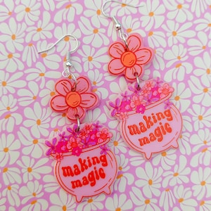 Può includere: Orecchini in acrilico rosa e arancio con un ciondolo a forma di fiore e un ciondolo a forma di calderone che dice "making magic".