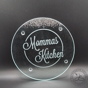 May include: A clear, round glass cutting board with the words "Mommas Kitchen" etched in a cursive font within a circle. The board has four small holes and a logo in the bottom right corner.