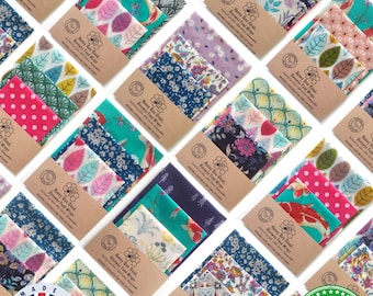 May include: A collection of folded beeswax food wraps featuring various colorful patterns, including floral, leaf, and polka dot designs. Each wrap is secured with a brown paper band labeled Honey Bee Good Beeswax Food Wraps.