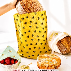 May include: A yellow beeswax bag with a black bee pattern containing a loaf of bread, a white cotton bread bag with a loaf of bread, a bowl of strawberries covered with a light green bee-patterned beeswax wrap, and an orange floral beeswax wrap.