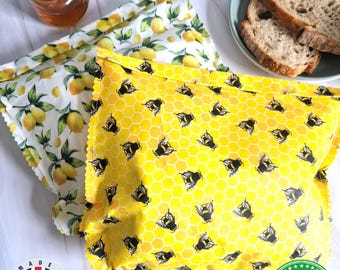 May include: Two rectangular beeswax food wraps. One wrap features a yellow honeycomb pattern with black bees, and the other features a white background with yellow lemons and green leaves. Text reads 100% HANDMADE IN THE UK.
