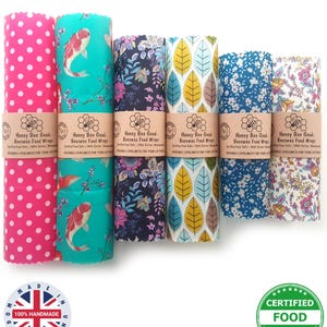 Könnte beinhalten: Sechs Rollen Honey Bee Good Bienenwachstücher mit verschiedenen Mustern, darunter rosa Punkte, Koi-Fische, Blumen und Blätter. Jede Rolle hat eine braune Papierbanderole mit dem Text Honey Bee Good Beeswax Food Wraps.